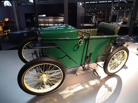 A green box-bodied electric car with cream wire wheels and a black seat in a dark exhibition hall.