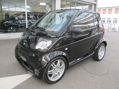 Glossy black two-seat convertible with Brabus Monoblock alloy wheels in a forecourt beside a showroom. Opens a larger view