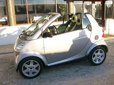 Silver open two-seater with green and black seats on cobbles in front of a restaurant’s windows in sunshine. Opens a larger view