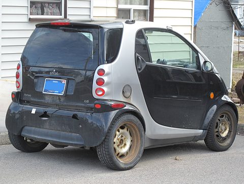 Rear of a black and silver two-seater with stacked tail lamps and a blue Ontario plate on gravel by a house. Opens a larger view