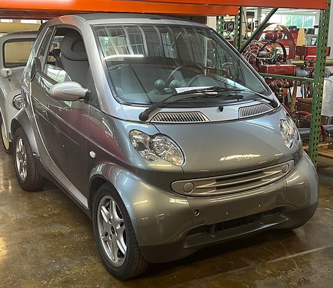 Metallic grey two-seat convertible with roof bars and alloy wheels on a museum floor, motorcycles behind. Opens a larger view