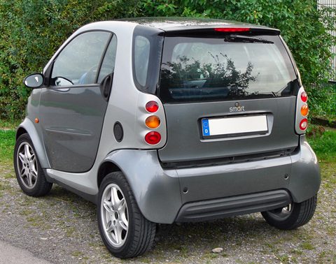 Rear of a grey and silver two-seater with stacked round tail lamps and a glass tailgate in front of shrubs. Opens a larger view