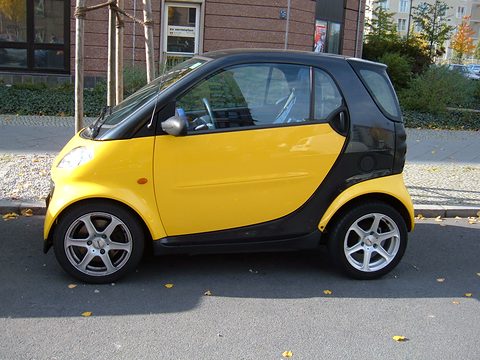 Side view of a short yellow two-seater with a black safety cell and alloy wheels at a kerb with autumn leaves.