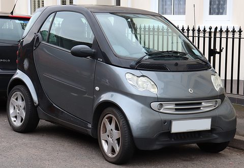 Dark grey two-seater with a chrome-barred grille and alloy wheels at a kerb beside iron railings. Opens a larger view