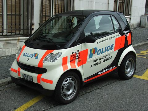 White and black two-seater with orange stripes and POLICE lettering parked on a street by a stone building. Opens a larger view
