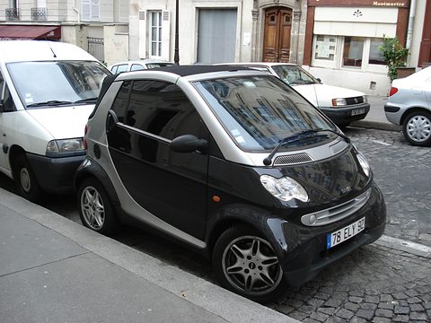 Black and silver two-seater with a Paris plate at the kerb between a white van and parked cars on a cobbled street. Opens a larger view