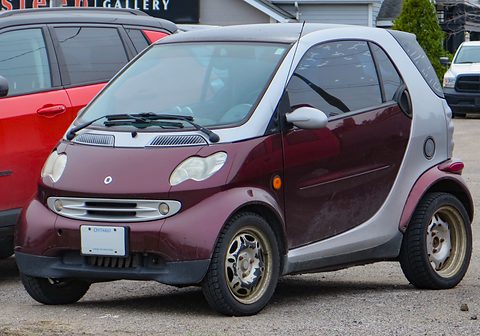 Dark red and silver two-seater with alloy wheels parked beside a red estate car on gravel. Opens a larger view