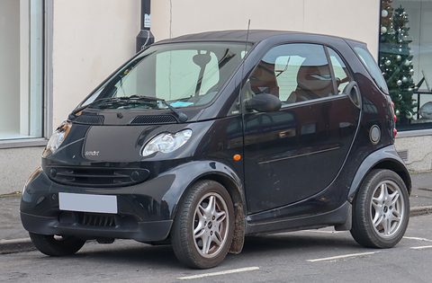 Black two-seater with steel-look wheels and a blanked plate parked by a shopfront with a Christmas tree. Opens a larger view