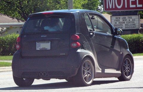 Rear of a dark grey two-seater with a twin central exhaust, parked beside a motel sign and hedges. Opens a larger view