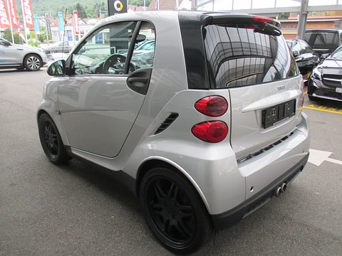 Rear of a silver two-seater with a black spoiler, round tail lamps, twin exhausts and black wheels. Opens a larger view