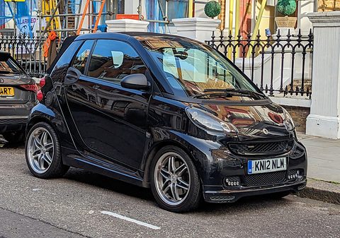 Black two-seater with Brabus alloy wheels and bodykit parked by iron railings and white pillars. Opens a larger view