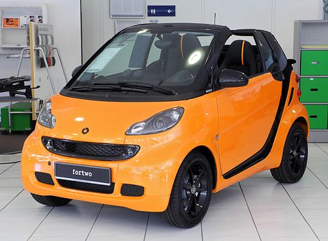 Bright orange open two-seater with black safety cell and black alloy wheels on a white tiled showroom floor. Opens a larger view