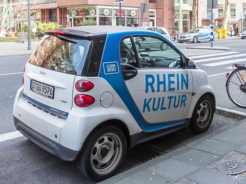 White and blue two-seater lettered RHEIN KULTUR parked at a kerb on a city street with brick buildings. Opens a larger view