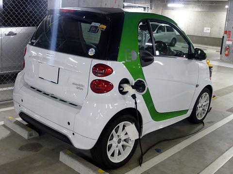 Rear of a white two-seater with a green frame, a charging cable plugged in where the fuel filler would be. Opens a larger view