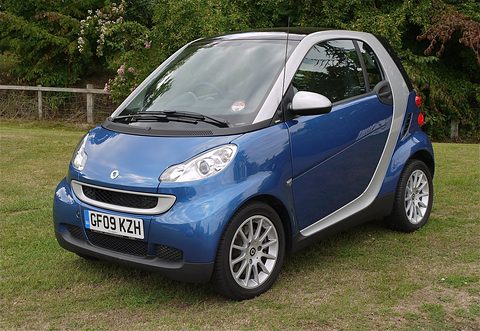 Blue two-seater with silver frame, round headlamps and alloy wheels on a grass verge beside bushes. Opens a larger view
