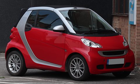 Red two-seater with a silver safety cell and alloy wheels on tarmac in front of a dark doorway. Opens a larger view