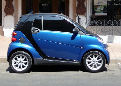 Side view of a blue and black two-seater with silver alloy wheels parked in front of a shop with iron grilles. Opens a larger view