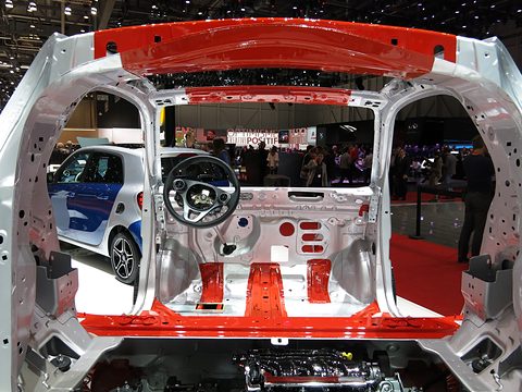 Silver unpainted car body frame with a red safety-cell outline, steering wheel and seat mounts on a show stand. Opens a larger view