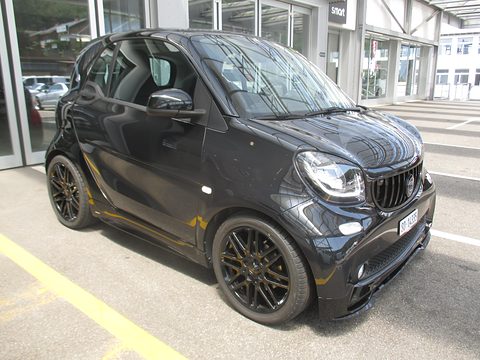 Glossy black two-seater with a Brabus grille and multi-spoke black wheels on a car park marked in yellow. Opens a larger view