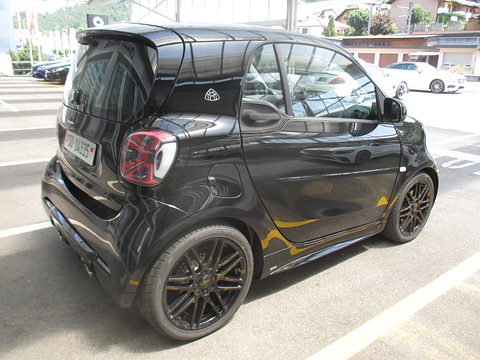 Rear of a black two-seater with a roof spoiler, red tail lamps and yellow decals in a parking bay. Opens a larger view