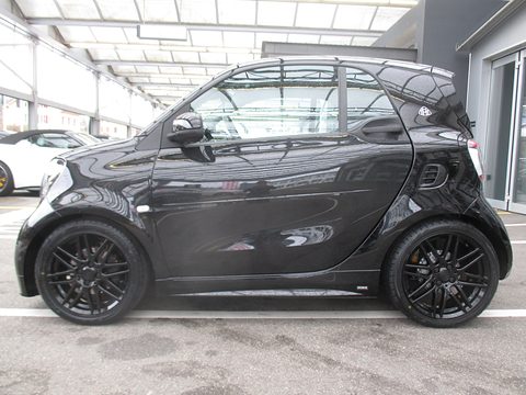 Side view of a glossy black two-seater with large black multi-spoke wheels under a covered car park. Opens a larger view
