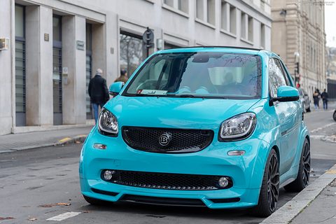 Turquoise two-seater with black Brabus grille and bodykit at a kerb in front of stone buildings. Opens a larger view
