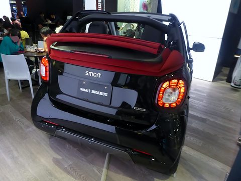 Rear of a black convertible two-seater with a folded red roof and round tail lamps on a wooden floor. Opens a larger view