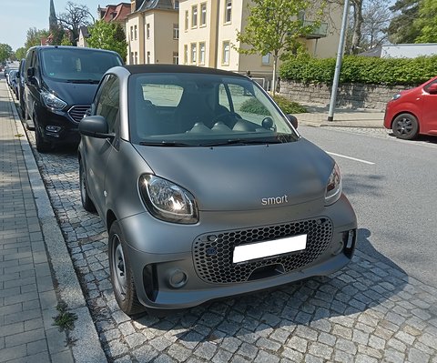 Matt grey electric two-seater with a honeycomb grille and blanked plate parked on cobbles beside a pavement. Opens a larger view