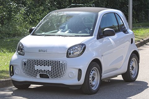 White electric two-seater with a honeycomb grille and black mirrors parked at a kerb beside grass.