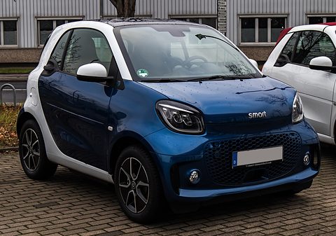 Blue electric two-seater with a white safety cell and black alloy wheels on brick paving. Opens a larger view