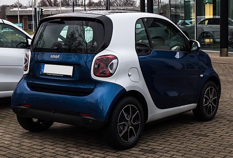 Rear of a blue electric two-seater with a white cell, large tail lamps and blanked plate on brick paving. Opens a larger view