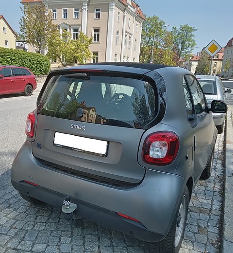 Rear of a matt grey electric two-seater with a tow bar and blanked plate on cobbles, houses behind. Opens a larger view