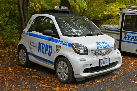 White two-seater with blue NYPD POLICE stripes and a roof light bar on autumn leaves beside a police van. Opens a larger view