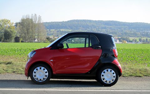 Side view of a red two-seater with a black safety cell and white wheel covers in front of fields and hills. Opens a larger view