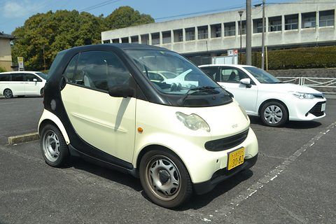 Cream two-seater with black frame and yellow kei plate in a car park beside a white hatchback and an office block. Opens a larger view
