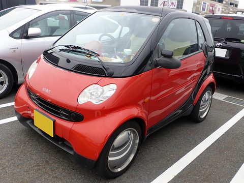 Red two-seater with a black frame, slim tyres and a yellow Japanese kei plate in a car park.