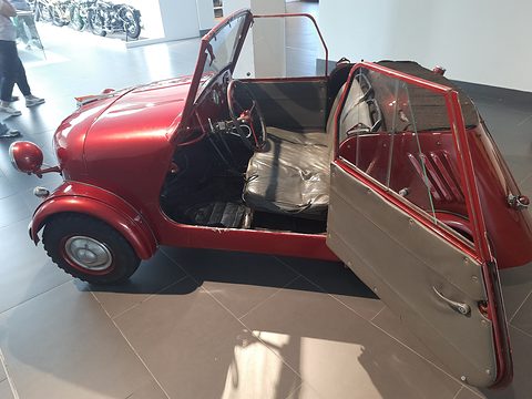 Red open microcar from the side, its door swung wide to show a black vinyl seat and hand-control levers.