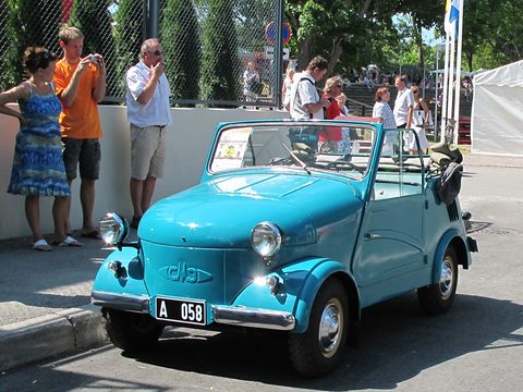 Turquoise open microcar with a plate A 058 parked in sunshine, people watching and photographing behind a fence. Opens a larger view