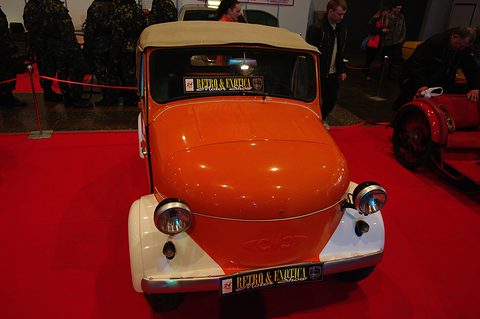 Orange microcar with cream wings and a beige hood, seen head-on on a red carpet, visitors behind. Opens a larger view