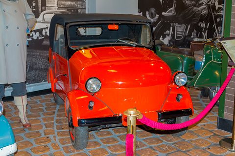 Bright orange-red microcar with a black hood, head-on behind a pink rope on a cobbled museum floor. Opens a larger view