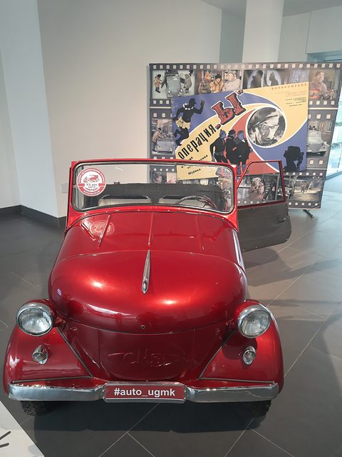 Front of a red open microcar with a pointed bonnet crease and two headlamps on its wings, a film poster behind. Opens a larger view