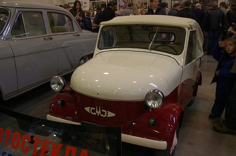 Red and white two-tone open microcar with a white hood up and the SMZ script on its nose, crowds behind. Opens a larger view