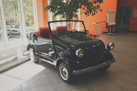 Black open microcar with red upholstered seats and chrome headlamps, on a tiled floor by tall windows. Opens a larger view