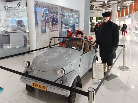 Pale grey open microcar with plate SMZ S-3A behind a rope in a shopping mall, costumed mannequins in and beside it. Opens a larger view
