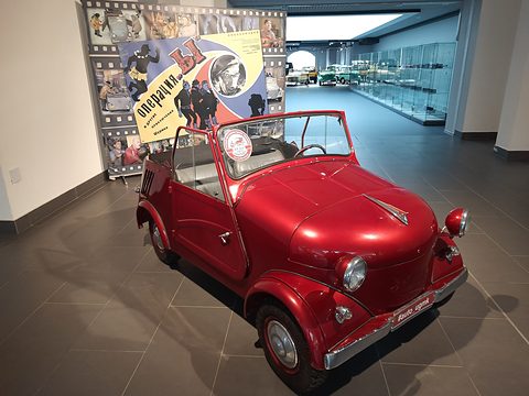 Glossy red open two-seater with round headlamps on its front wings, a colourful film poster on a stand behind.