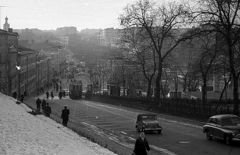 Black-and-white winter street scene: a snowy bank, bare trees, a tram in the distance and a small open car and saloon on the road.