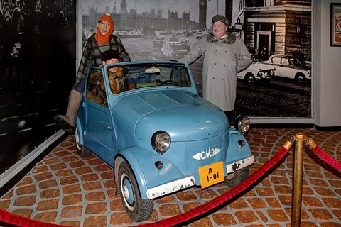 Pale blue open microcar with a number plate A 1-01, three costumed museum mannequins in and beside it before a street photomural. Opens a larger view