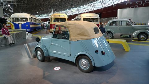 Pale blue microcar from the rear quarter, louvres on its tail and a beige folding hood, buses behind it in a long hall. Opens a larger view