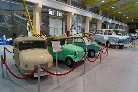 A line of small cars behind red ropes in a long museum hall: a beige three-wheeler, a green open car and a turquoise car half hidden beyond. Opens a larger view