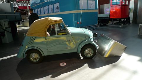 Side view of a small pale blue open car with a yellow hood, sunlight and shadow on the floor around it. Opens a larger view